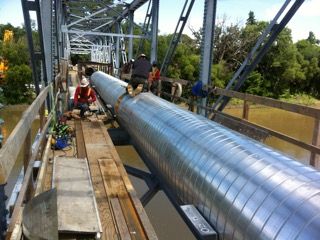 Crew installing a long insulated pipe along a truss bridge